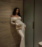 Woman in a white off-shoulder dress holding a clutch against a neutral wall.