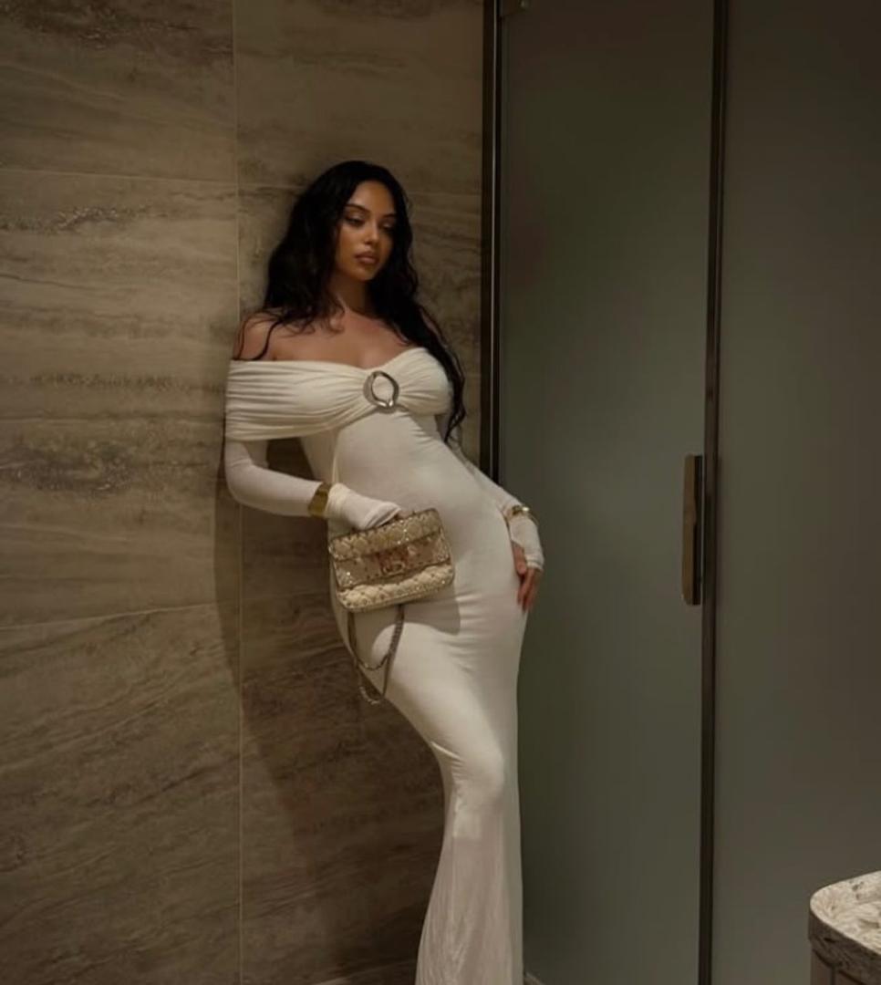 Woman in a white off-shoulder dress holding a clutch against a neutral wall.