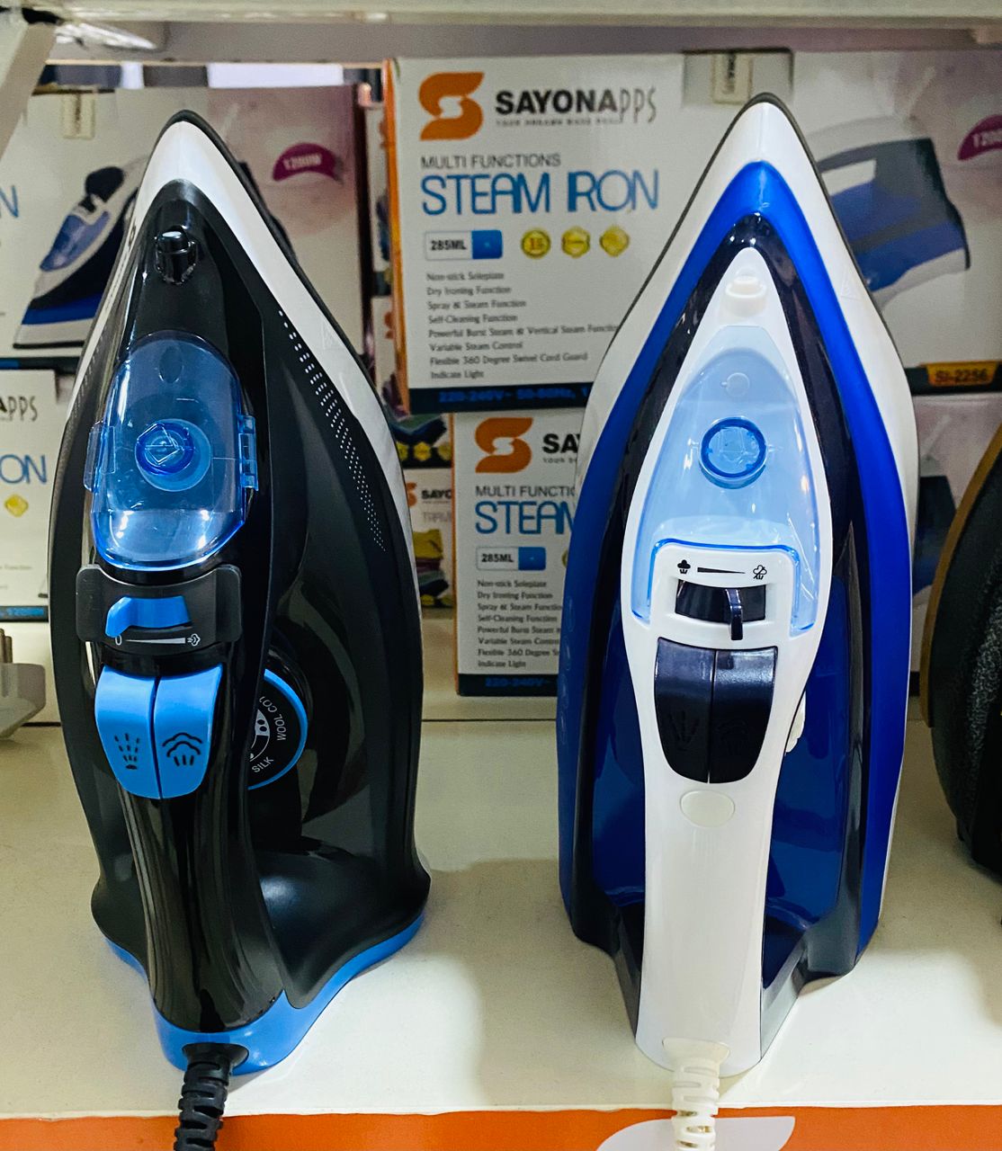 Two steam irons, one black and blue, one white and blue, on a shelf with promotional materials in the background.
